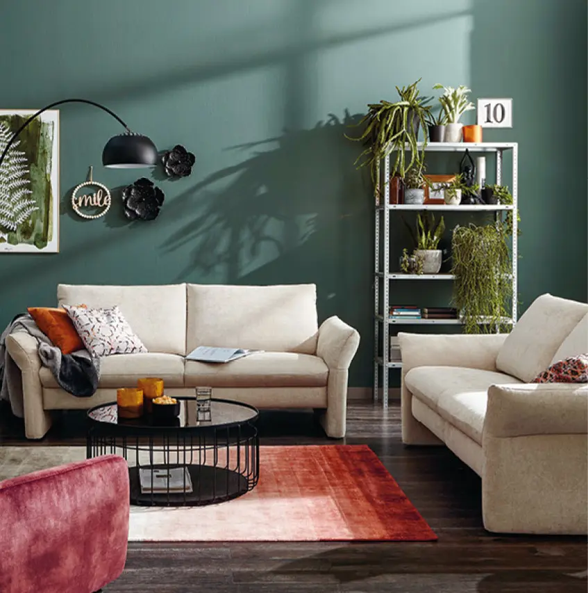 Zwei cremefarbene Sofas vor gruener Wand, Wanddeko links, schwarzer runder Couchtisch, Regal mit Pflanzen rechts, gemusterter Teppich